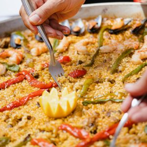 Close-up of hands serving seafood paella with fork, featuring shrimp and vegetables.